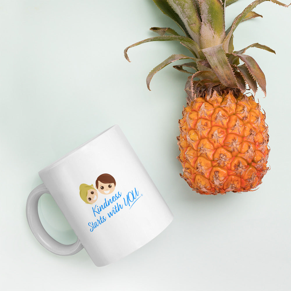 A white mug featuring the uplifting quote 'Kindness Starts with You" with a pineapple in the background.