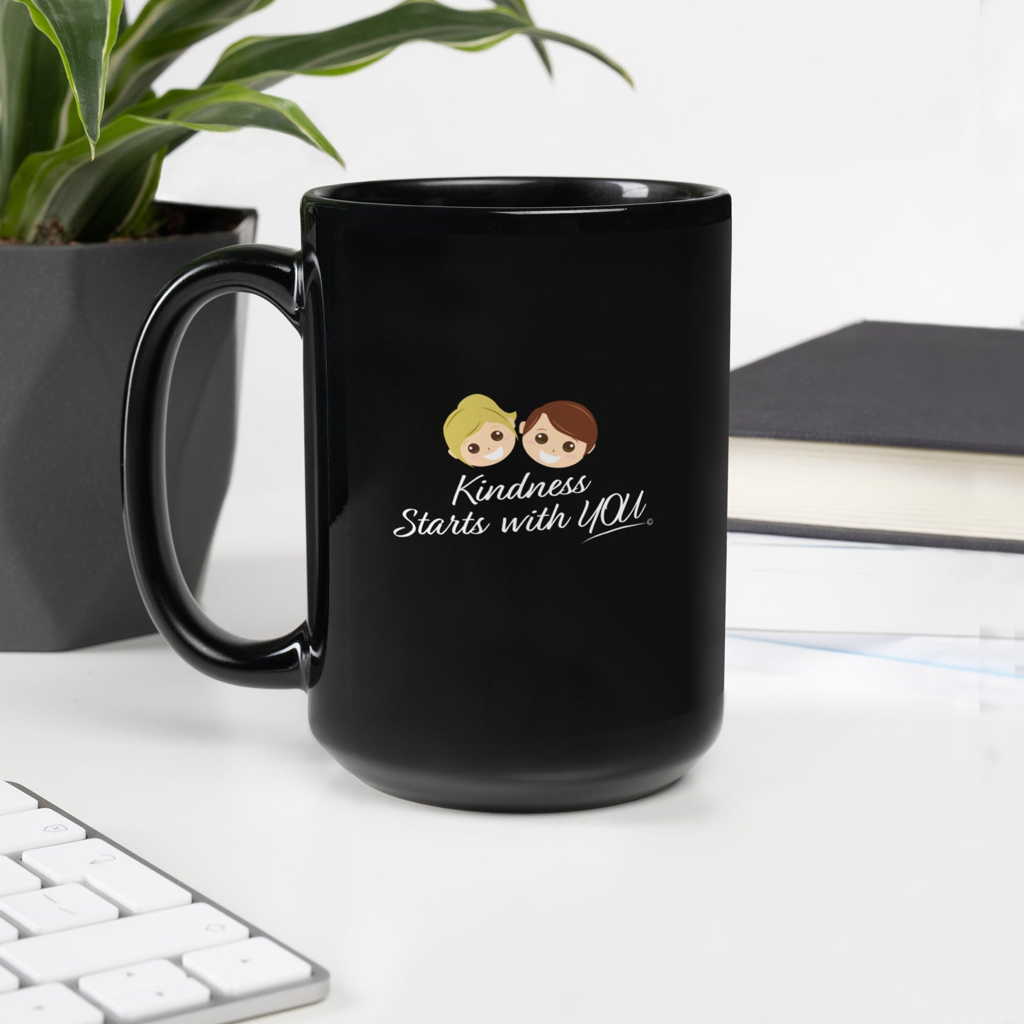 A 15-oz black mug featuring the uplifting quote 'Kindness Starts with You' with plant, keyboard and books in the background
